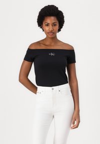 Zwarte off-shoulder top met korte mouwen, voorzien van een logo in het midden. Gecombineerd met witte hoge taille jeans. Gladde textuur, aansluitend ontwerp.