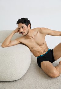 Schwarze Boxershorts mit einem blauen gemusterten Bund, glattem Stoff, figurbetontem Design und minimalen Nähten. Das Model posiert vor einem cremefarbenen Sofa.