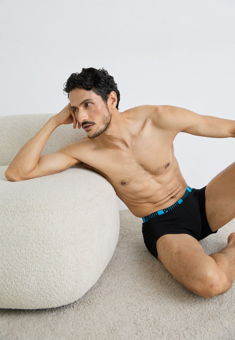 Schwarze Boxershorts mit einem blauen gemusterten Bund, glattem Stoff, figurbetontem Design und minimalen Nähten. Das Model posiert vor einem cremefarbenen Sofa.