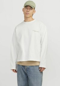 Sweatshirt alb scurt cu manșoni lungi și decolteu rotund, având un logo verde deschis. Asortat cu un top bej dedesubt și blugi din denim.