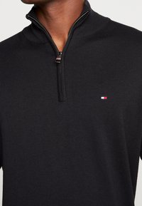 Maglione nero a collo zip in maglia con una texture a costine e un piccolo logo multicolore sul petto. Design aderente, maniche di lunghezza standard.