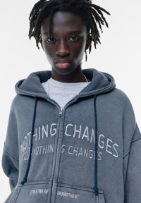 Grauer Reißverschluss-Hoodie mit auffälligem Sticktext, Kordeln und lockerer Passform. Über einem weißen T-Shirt getragen.