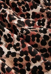 Tissu imprimé léopard avec des taches noires sur un fond beige, avec des accents rouges subtils. La texture semble douce et légère.
