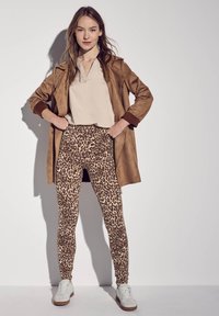 Ruskea mokkanahkainen pitkä takki, beige paita ja vaaleankeltainen leopardikuvioinen legginsit. Valkoiset lenkkarit ruskeilla yksityiskohdilla. Suorat pinnat ja istuvat muodot.