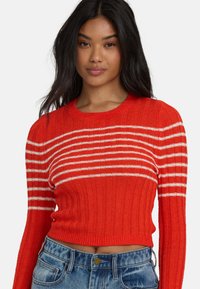 Czerwony sweter z ribbrowym splotem i białymi poziomymi paskami, z długimi rękawami i okrągłym dekoltem, zestawiony z dżinsami o wysokim stanie.
