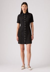 Zwarte denim jurk met korte mouwen, knoopsluiting aan de voorkant en twee borstzakken. Heeft zichtbare stiksels en een getailleerd silhouet.