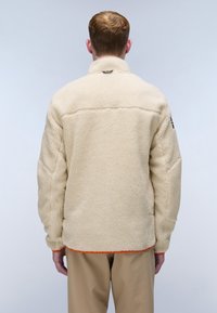 Cream-colored fleece jacket with a high collar, textured surface, and contrasting orange stitching at the hem. Features a discreet logo on the neck.