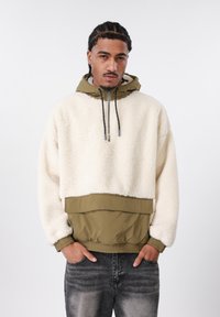 Kremo spalvos vilnos megztinis su alyvuogių žalumos apačios panelė ir kapišonas. Turi elastines rankogalius ir raištelį, taip pat kengūros kišenę.