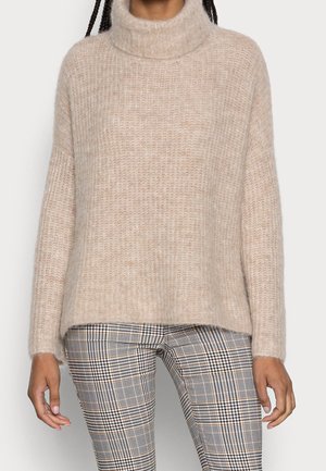 Pull à col roulé beige en tissu duveteux, associé à un pantalon ajusté à carreaux noirs, blancs et bruns, porté par une personne aux cheveux tressés.
