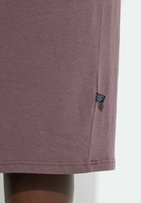 Primer plano del dobladillo de una prenda marrón-violácea con una pequeña etiqueta de tela negra en una pierna de piel oscura sobre un fondo pálido.