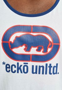 Witte tanktop met een blauw en rood grafisch ontwerp van een neushoorn binnen een logo, met de tekst "*eckō unltd." eronder.