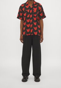 Uomo che indossa pantaloni larghi neri, scarpe nere e una maglietta nera a maniche corte con motivi di facce di diavolo rossi.