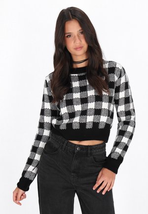 Kurzer Pullover mit einem schwarz-weißen Schachbrettmuster, langen Ärmeln mit schwarzen Bündchen und einer strukturierten Strickoberfläche. Kombiniert mit hochtaillierten schwarzen Hosen.
