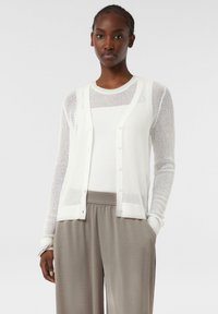 Weißer, gestrickter Cardigan mit Knopfverschluss und offenem Webmuster, über einem weißen, gerippten Oberteil getragen. Kombiniert mit hellgrauen, locker sitzenden Hosen.