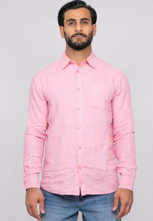 Homme aux cheveux foncés et à la barbe portant une chemise rose pâle boutonnée et un jean bleu, debout devant un fond clair uni.