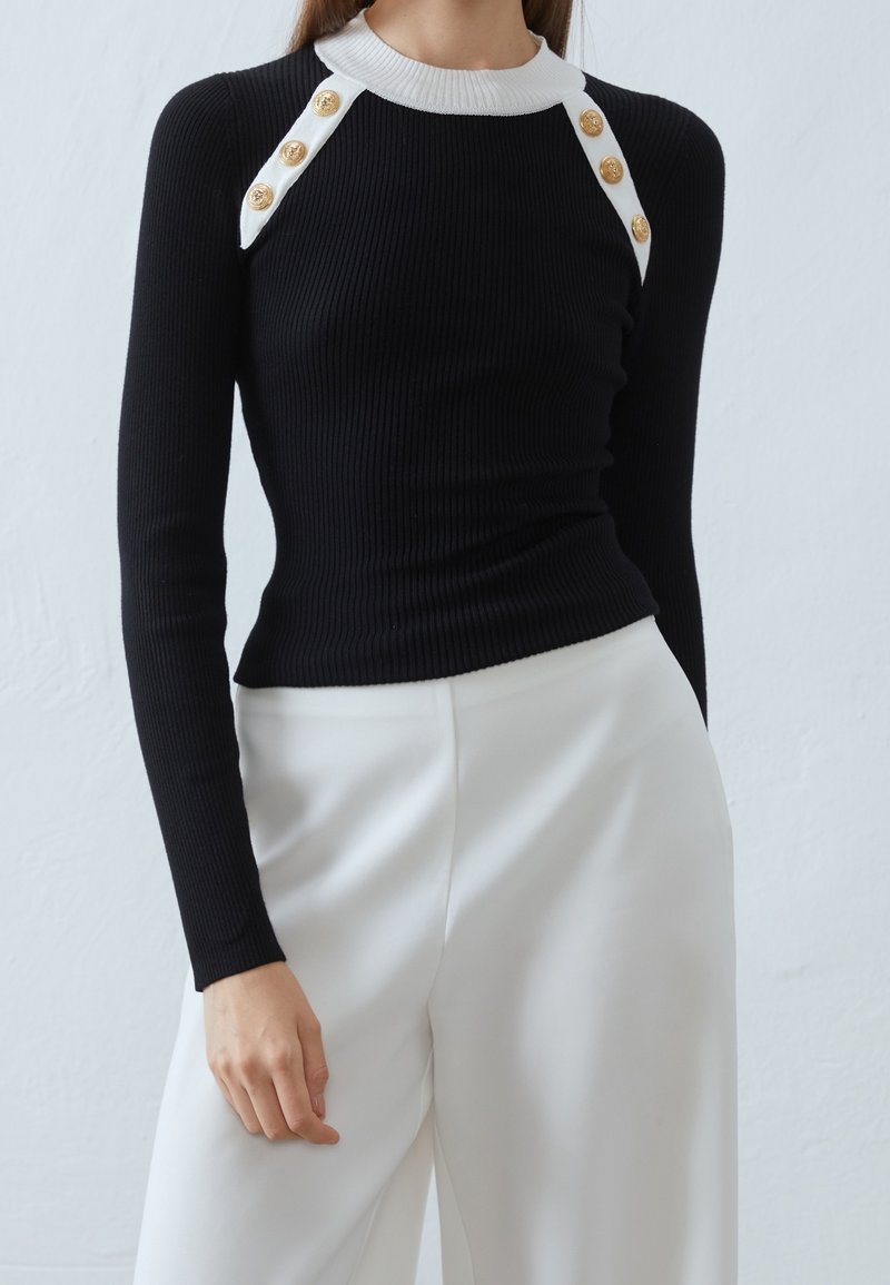 Haut noir en maille côtelée à manches longues avec col blanc et six boutons dorés, associé à un pantalon blanc taille haute à jambes larges.