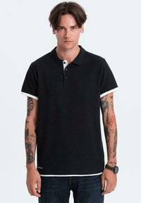 Polo shirt nera con colletto bianco e accenti bianchi sulle maniche. Maniche corte, presenta tre bottoni al collo e un logo ricamato sull'orlo.