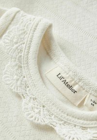 Tessuto bianco con motivo strutturato e bordo in pizzo sullo scollo. L'etichetta riporta "Lil' Atelier", indicando la taglia 1-2M. Materiale morbido, simile al cotone.