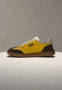 Baskets basses en cuir jaune, lacets blancs, empiècements marron sur l’avant et le talon, avec une semelle marron, présentées sur une surface claire.
