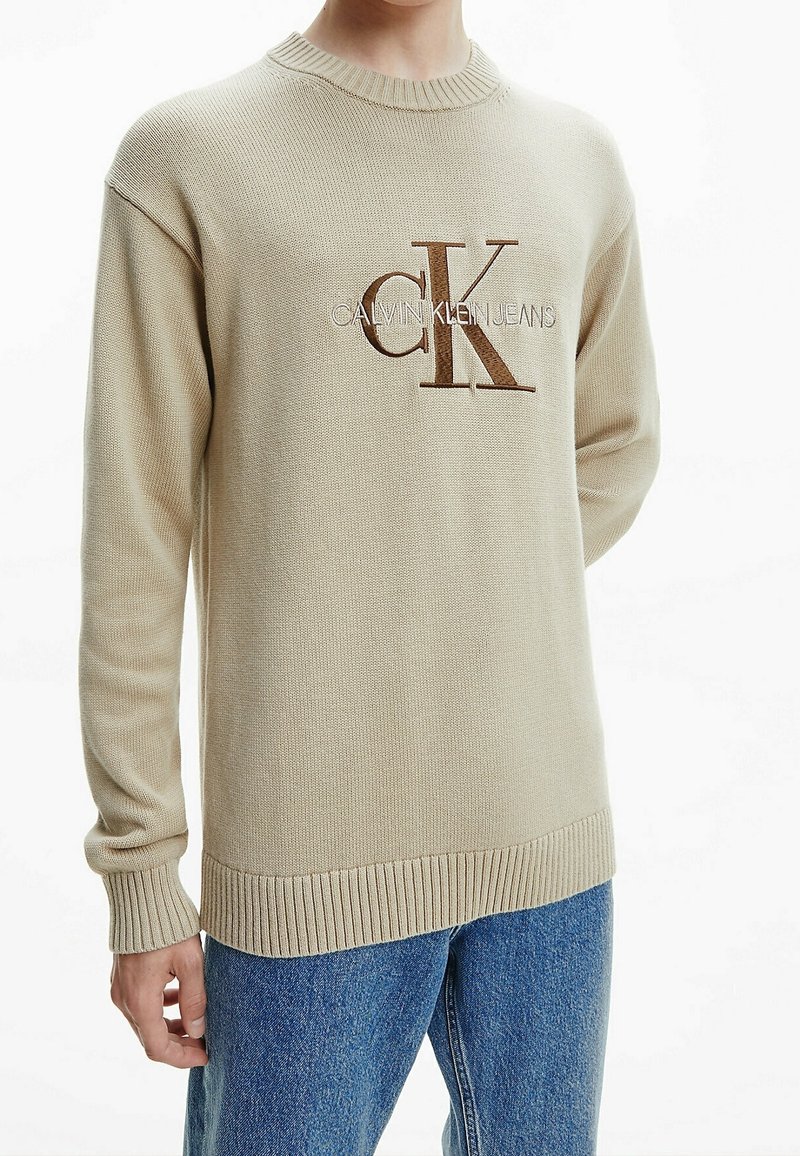 Pull en tricot beige avec poignets et ourlet côtelés, arborant un logo "CK" marron bien visible sur la poitrine. Col rond classique et coupe décontractée.