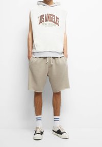 Personne portant un sweat à capuche blanc sans manches avec le texte « Los Angeles New Season », un short beige à cordon, des chaussettes blanches à rayures bleues et des baskets noires.
