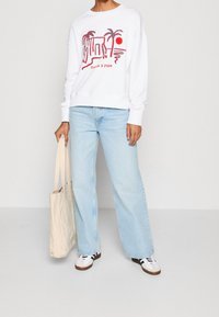 White sweatshirt with red graphic print of palm trees and sun, paired with light blue wide-leg jeans and white sneakers with black stripes.