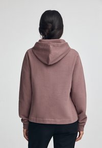 Sudadera color marrón con capucha, hecha de tejido suave. Presenta un ajuste holgado, puños acanalados y una textura lisa. Sin patrones ni elementos metálicos visibles.