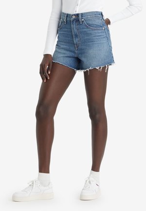 Personne portant un short en jean taille haute à ourlet effiloché, un haut blanc à manches longues côtelé, des baskets blanches et des chaussettes blanches montantes, debout avec la main dans la poche.
