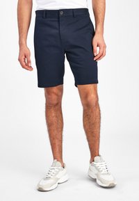 Pantaloni scurți de culoare bleumarin, dintr-un material neted, cu închidere frontală cu nasture și buzunare laterale; asortați cu adidași de culoare deschisă.