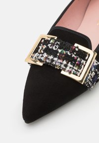 Chaussure plate noire à bout pointu avec bride en tweed multicolore et boucle rectangulaire dorée sur fond blanc.