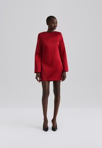 Red satin mini dress with long sleeves and a square neckline. Smooth texture and structured silhouette. Worn with black pointed heels.