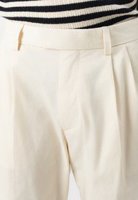 Pantalones de color beige claro confeccionados en algodón, con un diseño de frente plano, pliegues, bolsillos laterales y una textura suave. Cintura con trabillas para cinturón.