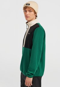 Groene fleece pullover met zwarte en crèmekleurige accenten, ritskraag, fitted manchetten en subtiele branding op de borst en de beanie.