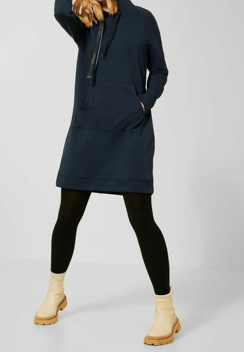 Marinblå hoodieklänning med en framficka, har en dragsko i huvan och är parat med svarta tights och beige ankelstövlar.