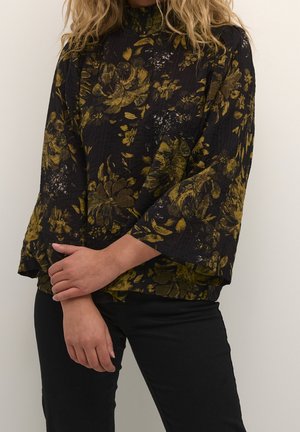 Kobieta ubrana w czarną bluzkę z dużymi żółtymi kwiatowymi wzorami i czarne spodnie, stojąca z rękami skrzyżowanymi na nadgarstkach.