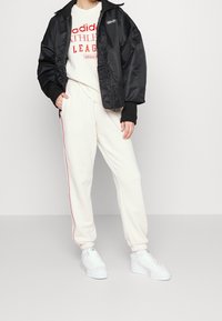 Svart sating bomberjacka, vit grafisk sweatshirt, krämfärgade joggers med röd sidstrimma och vita sneakers. Texturerade tyger och sportig design.