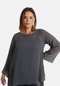 Blusa trasparente grigio scuro con maniche lunghe, caratterizzata da accenti luccicanti ai polsini. Tessuto morbido e scollatura rotonda. Design leggero e morbido.