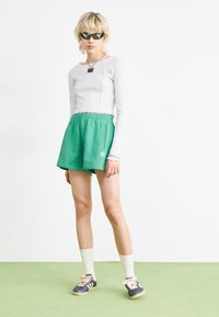 Πράσινες αθλητικές βερμούδες με ελαστική μέση, συνδυασμένες με λευκή μακρυμάνικη μπλούζα με ραβδώσεις. Μαύρα κομψά γυαλιά ηλίου και γκρι παπούτσια με κρέμ λεπτομέρειες.