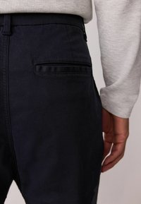 Tmavomodré nohavice s hladkým povrchom a jednou zadnou vreckom. Čiastočne je vidieť šedú, textúrovanú košeľu s dlhým rukávom.