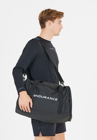Musta duffel-laukku, jossa on valkoinen "ENDURANCE"-logo. Valmistettu kestävästä kankaasta, jossa on kaksi kahvaa ja irrotettava olkahihna.