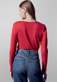 Rode langemouwen top gecombineerd met hooggesneden blauwe jeans. De top heeft een ronde halslijn; de jeans hebben twee achterzakken en een gestructureerde afwerking.