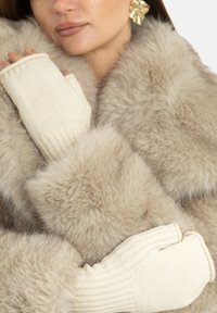 Beżowy płaszcz z futra o miękkiej teksturze; z kremowymi, bezpalczastymi rękawiczkami z prążkowanego materiału. Zdjęcie z bliska podkreślające ciepło i warstwowość.