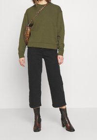 Sudadera verde oliva con puños y dobladillo acanalados, combinada con jeans de pierna ancha negros. Bolso cruzado con estampado de serpiente en contraste y botines de tacón negro.