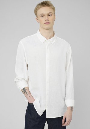 Jeune homme aux cheveux blonds courts, portant une chemise blanche à manches longues boutonnée et un pantalon foncé, debout avec une main dans la poche.