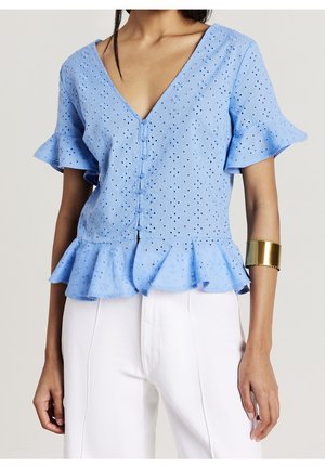 Femme vêtue d'un chemisier bleu clair en broderie anglaise avec manches et ourlet à volants, associé à un pantalon blanc et un large bracelet manchette en or au bras gauche.
