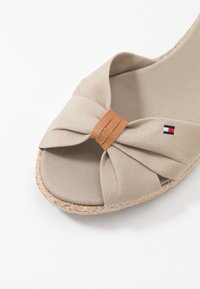 Sandalia espadrille de tela beige con un lazo retorcido y un acento de cuero en la puntera, suela de yute y un pequeño logo rojo, blanco y azul en la correa.