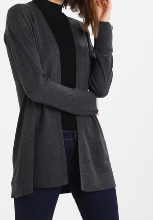 Femme portant un cardigan gris foncé ouvert sur un col roulé noir et un jean bleu foncé, posant avec une main près du visage.