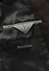 Zwarte jasinterieur met satijnen voering en een gestreepte zak. Het label leest "Matinique" in wit op een zwarte stoffen patch.