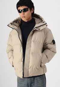 Beige pufferjack met een rits aan de voorkant, een capuchon en een textuurstof. Voorzien van zijzakken en een zwart logo-opdruk op de mouw.