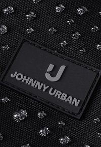 Close-up af sort stof med vanddråber og en sort rektangulær label med teksten "Johnny Urban" i grå bogstaver.